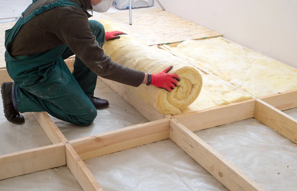 Ouvrier en salopette verte installant de l'isolant thermique en laine minérale entre solives de plancher en bois