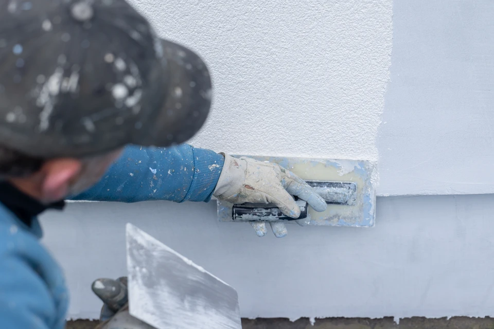 Vue d'un chantier de rénovation de maison montrant un artisan en tenue professionnelle (gilet haute-visibilité ou costume de travail) observant ou inspectant à la fois une façade en cours de ravalement (avec échafaudage et enduit frais visible) et l'accès à l'intérieur du bâtiment où sont visibles des travaux de rénovation intérieure (par exemple, zone de peinture ou isolation intérieure). L'image doit transmettre une impression de prise en charge globale et de continuité entre travaux extérieurs et intérieurs, avec un seul professionnel responsable de l'ensemble.