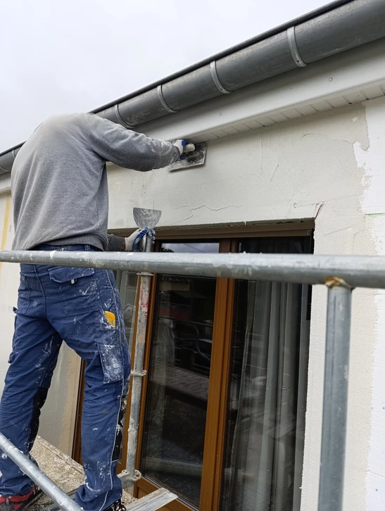 Ouvrier du bâtiment en vêtements de travail et casque blanc appliquant un enduit ou peinture blanche sur façade extérieure depuis échafaudage métallique