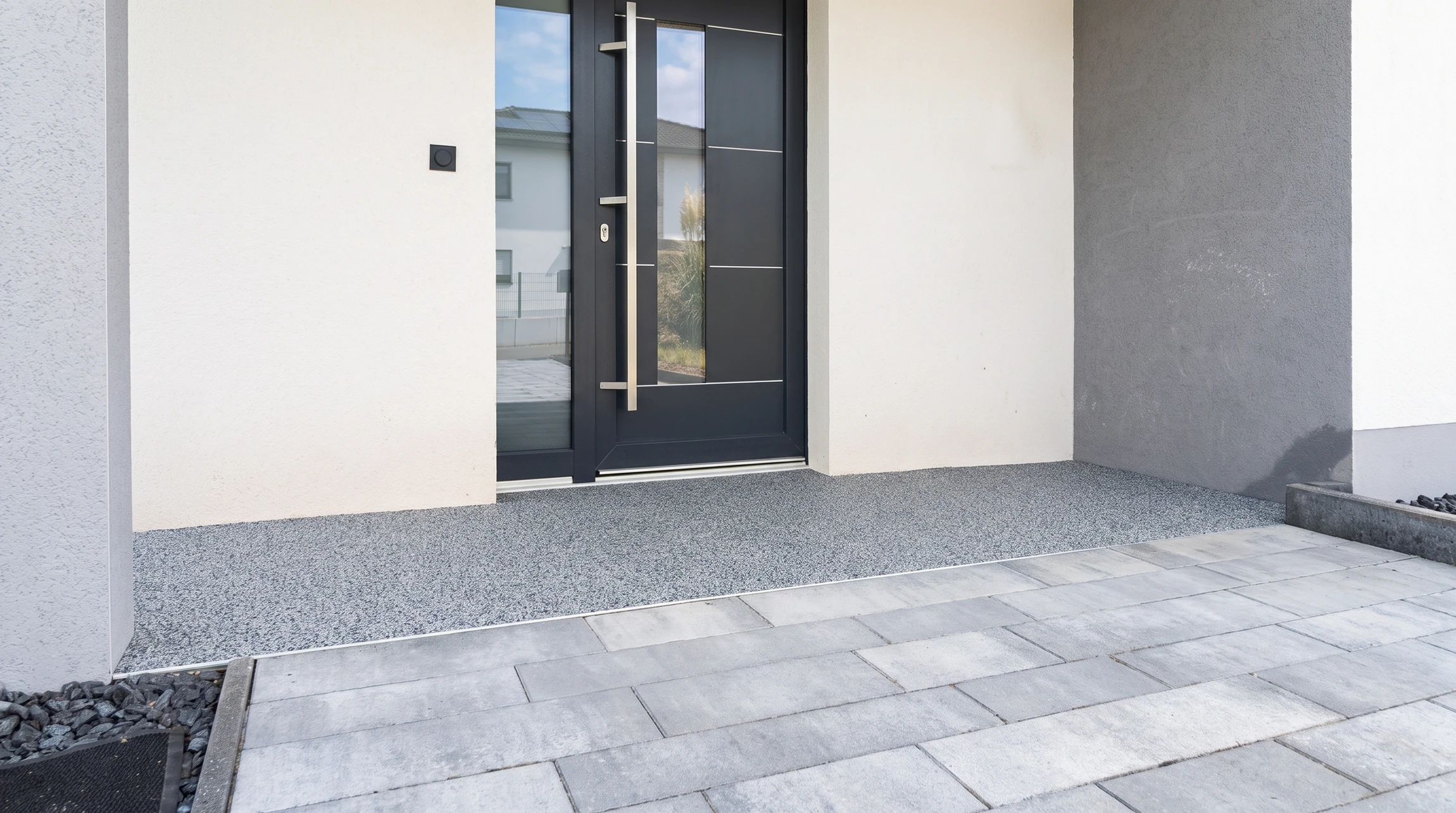 Photographie d'une terrasse extérieure moderne récemment rénovée, mettant en évidence un revêtement de moquette de pierre (agrégats colorés et résine) couvrant toute la surface au sol. La texture gravillonnée et brillante du matériau doit être clairement visible et bien mise en lumière. La terrasse peut être aménagée avec quelques éléments sobres (une chaise simple, une plante) pour donner du contexte, mais l'accent doit rester sur la qualité et l'esthétique du revêtement de sol en résine.