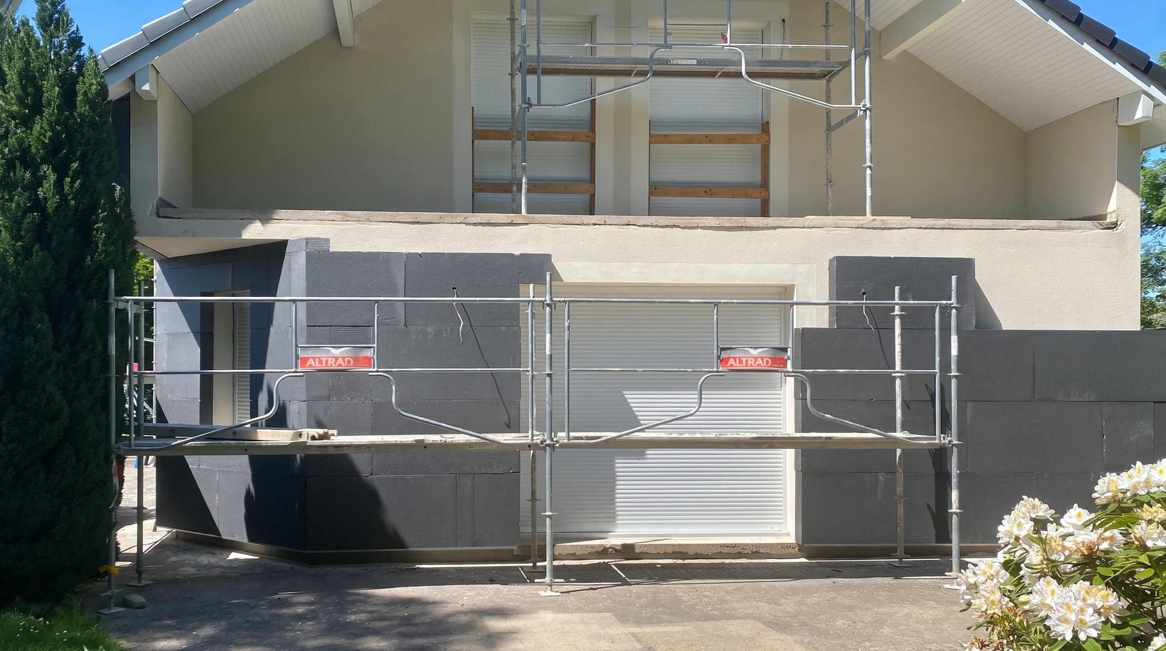 Photographie réaliste d'un ouvrier en tenue de travail professionnelle appliquant une couche d'isolation thermique (panneau isolant ou enduit de finition) sur la façade extérieure d'une maison de résidence. La façade doit clairement montrer un bâtiment résidentiel en cours de rénovation extérieure, avec des éléments visibles d'isolation en place (panneaux blancs ou beiges) et l'ouvrier utilisant les outils appropriés (taloche, spatule) pour l'application. La scène doit transmettre une atmosphère professionnelle et de qualité, avec une maison rénovée et soignée en arrière-plan.